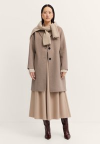 Beige Wollmantel mit strukturierter Silhouette, ausgestattet mit schwarzen Knöpfen und einem cremefarbenen gerippten Schal. Getragen über einem langen, fließenden beigen Rock.