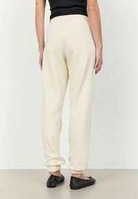 Beiga sweatpants med elastiskt midjeband och cuffs, med en mjuk struktur. Bärs med svarta platta skor, visad från baksidan.