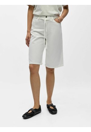 Personne portant un short blanc jusqu'aux genoux avec une ceinture nouée, une chemise gris clair, des chaussures plates noires, et un bracelet argenté au poignet gauche.