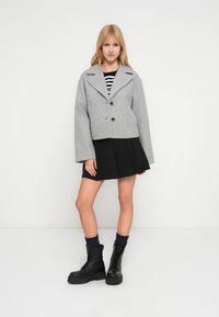 Γκρι κοντό μπουφάν με μεγάλο γιακά, δύο κουμπιά και φαρδιά μανίκια, συνδυασμένο με μαύρη φούστα και μαύρες μπότες με κορδόνια. Φαίνεται ριγέ τοπ.