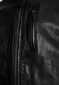 Svart läderjacka med en slät yta, som har en framträdande dragkedjedetalj och en liten diagonalt placerad dragkedjeficka på bröstet.