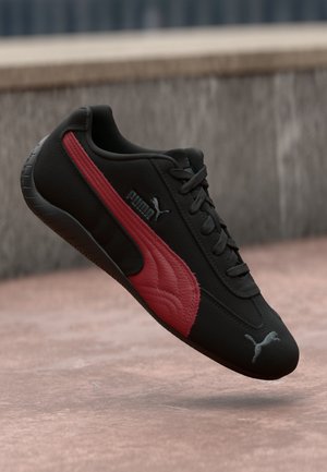 Zwarte sportschoen met een rood, textuur detail aan de zijkant, vetersluiting aan de voorkant en het Puma-logo. Slank ontwerp met een robuuste rubberen zool.