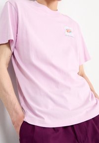 Osoba w jasnoróżowej koszulce z napisem i logo "United Youth", w zestawieniu z ciemnofioletowymi spodniami, z rękami w kieszeniach, na białym tle.