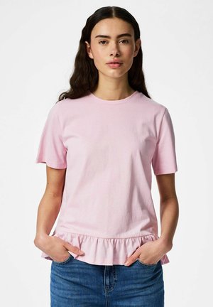 Ženska z dolgimi temnimi lasmi, oblečena v svetlo roza kratkoročni vrh z volančastim robom in modre kavbojke, roke v žepih, nevtralen izraz.