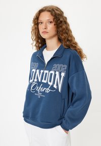 Sudadera de forro polar azul con medio cierre, que presenta un texto gráfico blanco "LONDON Oxford" y los años 1998, 2002. Puños y dobladillo acanalados, corte relajado.