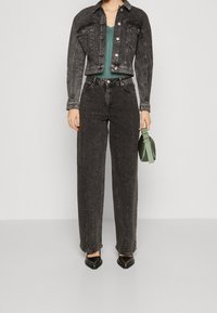 Žena oblečená v černé džínové bundě a odpovídajících džínách s vysokým pasem a širokými nohavicemi s drobnými bílými skvrnami, držící zelenou kabelku.