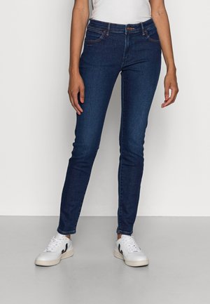 Persona con vaqueros ajustados azul oscuro y zapatillas blancas, de pie contra un fondo liso y claro.