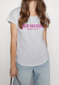 Svetle šedé tričko s krátkým rukávem, na kterém je tmavě růžové logo "TRUE RELIGION" a text "BRAND JEANS". Volný střih, nošeno s modrými širokými džíny.