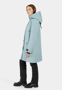 Ljusblå vattentät parka-jacka med frontdragkedja, stora fickor, justerbar huva och svarta detaljer; kombinerad med svarta jeans och stövlar.