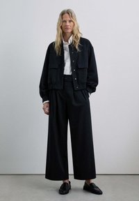 Mujer con cabello largo y rubio, vestida con una camisa blanca, una chaqueta negra holgada, pantalones anchos negros y mocasines negros, de pie contra una pared clara.