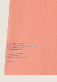 Camiseta de algodón color durazno con texto plateado que contiene frases motivacionales y un logo en la esquina inferior izquierda. Manga corta, cuello redondo.