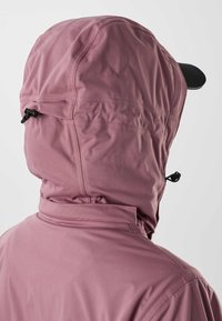 Personne portant une veste à capuche mauve avec une casquette de baseball noire sous la capuche, vue de dos sur un fond uni.