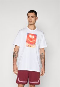 Jonge man met kort donker haar, gekleed in een wit t-shirt met een rode smiley-gezicht grafiek en bordeauxrode sportshorts met roze afwerking.