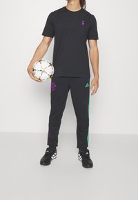 Černé sportovní tričko s fialovým logem, černé kalhoty s zelenými a fialovými bočními pruhy a černé fotbalové boty, držící barevný fotbal.