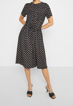 Femme portant une robe midi noire avec des motifs circulaires blancs, à manches courtes, avec une ceinture à la taille, et des mules ouvertes à talons noirs, se tenant les mains derrière le dos.