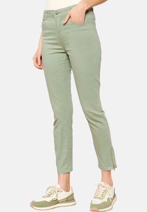 Smaltsiddende lysegrønne bukser med lommer foran og lynlåse ved anklerne, båret med beige og grønne sneakers. Én hånd i lommen, underkroppen vist.