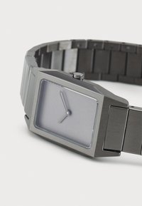 Sølvtonet rektangulær klokke med mat finish, minimalistisk design og grå urskive. Har en enkel time- og minutt hånd. Metallslenke.