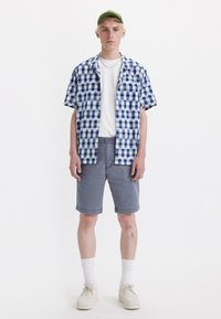 Camisa de cuadros azul y blanca de manga corta, camiseta blanca debajo, pantalones cortos grises, calcetines blancos y zapatos beige, de pie frente a un fondo blanco.