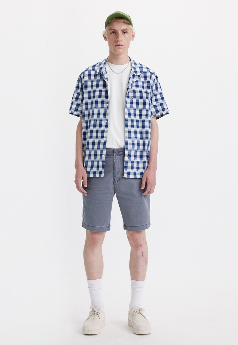 Camisa de cuadros azul y blanca de manga corta, camiseta blanca debajo, pantalones cortos grises, calcetines blancos y zapatos beige, de pie frente a un fondo blanco.