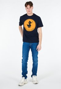 T-shirt blu navy con una grafica circolare arancione raffigurante un'anatra e il testo "SALVA L'ANATRA". Abbinato a jeans blu denim e sneakers chiare.