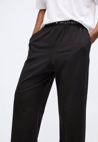 Pantalons noirs avec une taille élastique arborant un logo "Tommy Hilfiger". Le tissu semble lisse, avec des poches latérales et une coupe décontractée.
