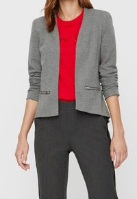 Grå strikket blazer med lange ærmer, åben front og to lynlåslommer. Båret over en rød t-shirt og parret med grå bukser.