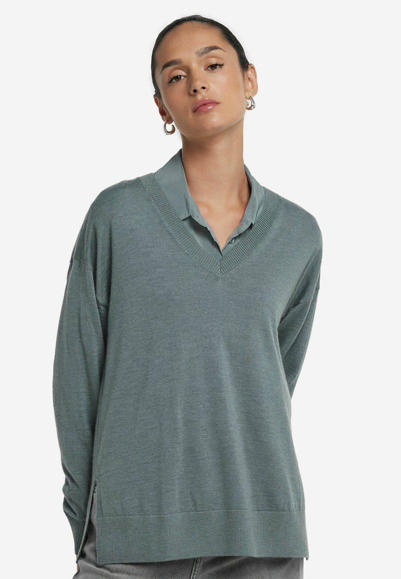 Kvinna med mörkt hår i en låg knut, iförd silvriga hoopörhängen, en ljusgrön skjorta med krage och en överdimensionerad dämpad grön V-ringad tröja.