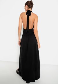 Melnā halter-stila maksi kleita ar izsmalcinātu vidukli, kas piedāvā garas saites ap kaklu un plūstošu svārku. Gluda auduma, minimālas detaļas.