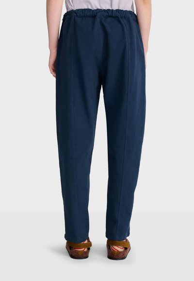 Personne portant un pantalon bleu marine à taille élastique et des sandales marron, debout devant un fond clair uni, vue de dos.