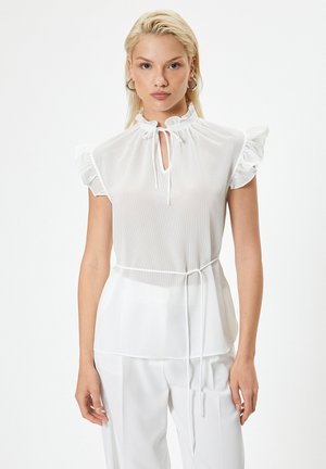 Joven mujer con cabello rubio que lleva una blusa blanca sin mangas con volantes y lazo en el cuello y pantalones blancos sobre un fondo blanco.