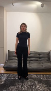 Moteris su šviesiai rudais plaukais, dėvinti juodą marškinius ir kelnes, stovi ant raštuoto kilimo priešais pilką odinę sofą minimalistiniame kambaryje.