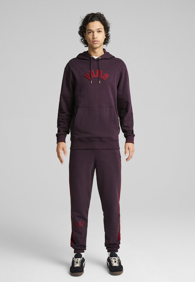 Bordeauxfarbener Hoodie mit Logodetail und Kordelzügen, kombiniert mit passenden bordeauxfarbenen Jogginghosen mit Seitenstreifen und Branding. Das Material wirkt weich.