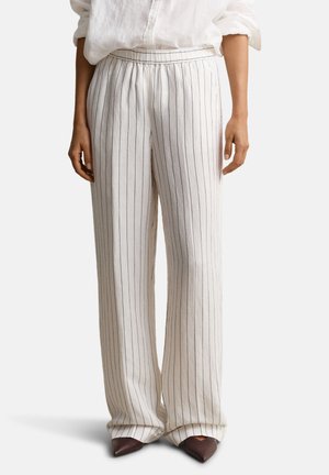 Frau trägt weiße Hosen mit weitem Bein und dünnen vertikalen schwarzen Streifen sowie braune spitze Schuhe, kombiniert mit einem weißen Langarmshirt.