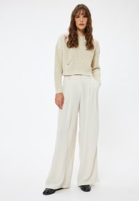Crème gebreide sweater met een cropped ontwerp en ribbelstructuur, gecombineerd met losse, high-waisted, lichtgekleurde plooibroeken. Zwarte schoenen.