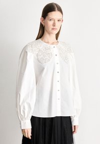 Biela blúzka s veľkým, prepracovane vyšívaným golierom so striebornými akcentmi, dlhými nafúknutými rukávmi a zapínaním na gombíky vpredu.