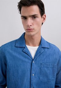 Camisa de denim en azul medio con cuello, cierre de botones y un bolsillo en el pecho. Textura suave, corte relajado y detalle de costura visible.