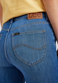 Gros plan d’une personne portant un jean bleu Lee et un haut jaune, mettant l’accent sur la poche arrière et le patch de la marque Lee.