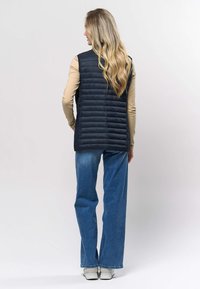 Črna puhasta telovnik z navpičnim vzorcem prešite tkanine, kombiniran s svetlo bež majico z dolgimi rokavi in modrimi jeans hlačami širokih nogavic. Gladek tekstura, sproščen kroj.