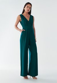 Teal ærmeløs jumpsuit med dyb V-udskæring, defineret talje, brede ben og sidelommer. Båret med strappy guldhæle. Glat stofstruktur.
