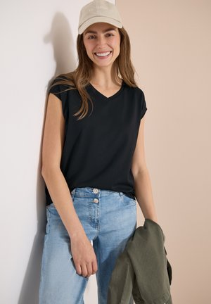 Smilende kvinde iført beige kasket, sort ærmeløs top, lyseblå jeans, der holder en olivengrøn jakke, stående mod en beige og hvid væg.