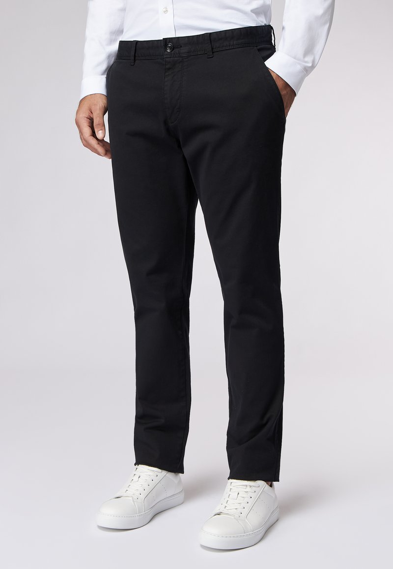 Schwarze Baumwollhosen mit schmaler Passform, ausgestattet mit Seitentaschen und einer glatten Textur. Getragen mit weißen Sneakern für den Kontrast.