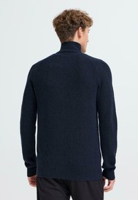 Námořnická modrá pletená svetr s vysokým límcem, dlouhými rukávy a volným střihem. Textura je silná s jemnými variacemi v odstínech.