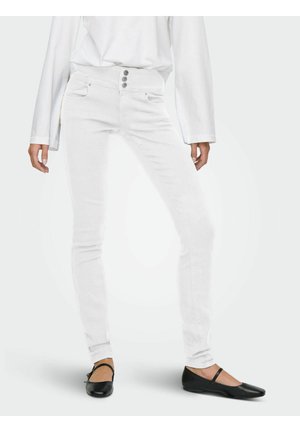 Biele úzke rifle so tromi prednými gombíkmi nosené s čiernymi balerínami Mary Jane a bielym tričkom s dlhým rukávom, zobrazené od pásu po nohy.