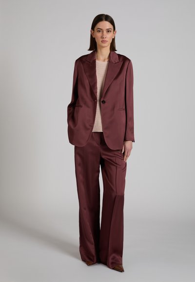 Mujer con una chaqueta oversized de color burdeos brillante y pantalones de pierna ancha a juego, junto con un top de punto beige, posando frente a un fondo gris liso.