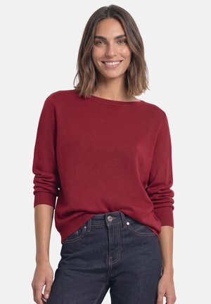 Kvinde med skulderlangt brunt hår iført en rød langærmet sweater og mørkeblå jeans, smilende mod en ensfarvet baggrund.