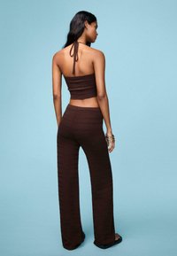 Smeđi pleteni crop top s halter izrezom i širokim hlačama koje odgovaraju. Tekstura sadrži horizontalne pruge. Nosila ga je manekenka protiv plave pozadine.