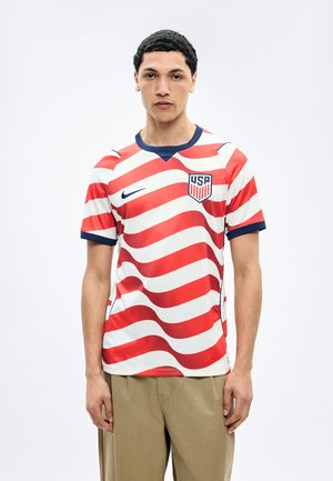 Jaunas vyras dėvi smėlio spalvos kelnes ir trumpomis rankovėmis JAV futbolo marškinėlį su raudonomis ir baltomis banguotomis juostelėmis, stovintis priešais paprastą baltą foną.