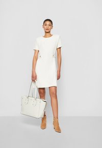 Witte jurk met korte mouwen, een aansluitende taille en twee decoratieve knopen, gecombineerd met een witte gestructureerde handtas en tan enkellaarsjes.