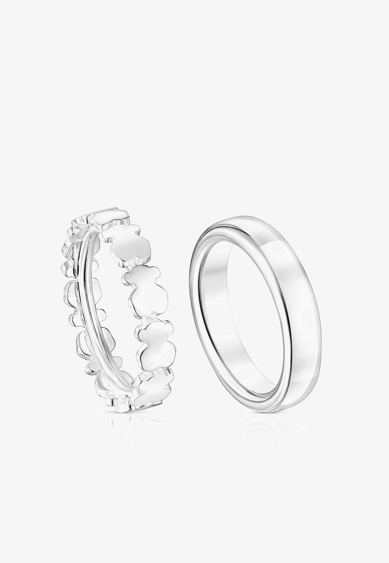 Dos anillos de plata uno al lado del otro, uno con una banda sólida y lisa y el otro con un patrón entrelazado de formas de osos de peluche.
