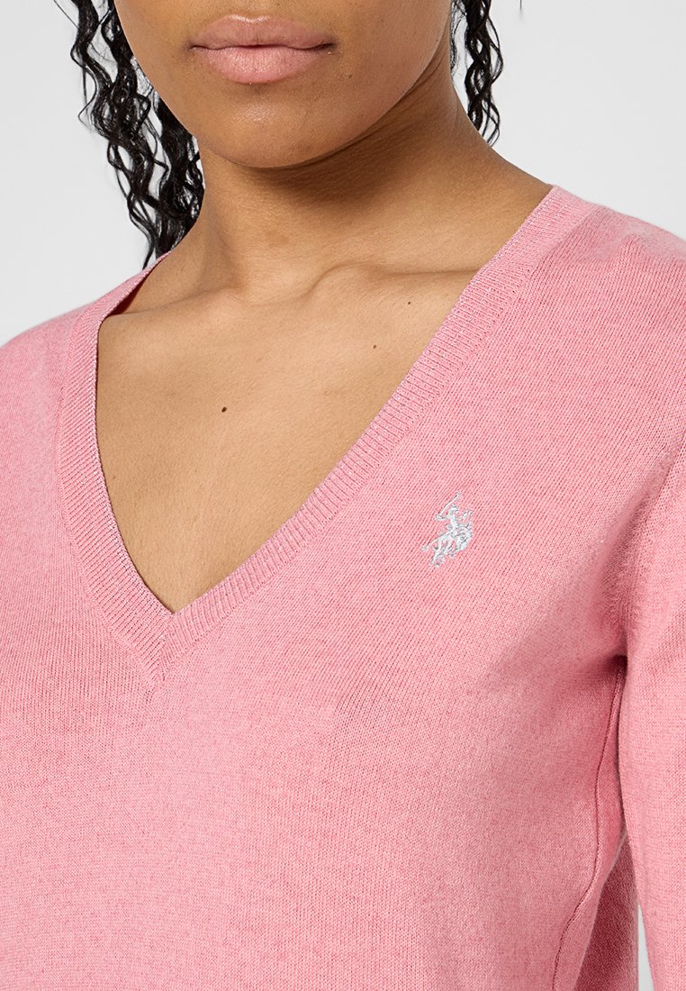 Donna che indossa un maglione rosa chiaro a scollo a V con un piccolo logo ricamato bianco sul lato sinistro del petto e capelli ricci raccolti.
